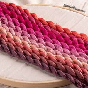 Pode incluir: Fios de bordar em tons de rosa, vermelho e laranja, dispostos diagonalmente em um bastidor de madeira. As cores variam do roxo escuro ao laranja brilhante. O tecido é bege claro.
