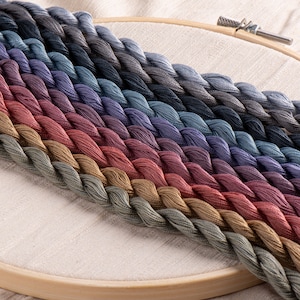 Puede incluir: Una colección de carretes de hilo de bordar en un degradado de colores apagados, que van desde el azul oscuro hasta el marrón claro. Los carretes están dispuestos en forma de abanico sobre un aro de bordado de madera.