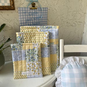 Puede incluir: Cuatro bolsas de tela acolchadas en un patrón floral amarillo y azul. Las bolsas están apiladas en una mesa blanca. Las bolsas están etiquetadas con "TheChaveliEdit".