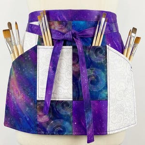 Pode incluir: Um avental de artista roxo e com tema de galáxia com bolsos cheios de pincéis. O avental tem um design de cintura com laço e é feito de tecido com um padrão de galáxia rodopiante e painéis brancos com um design de redemoinho.