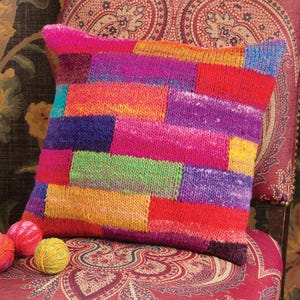 May include: A colorful knitted pillow with a patchwork design in shades of pink, purple, green, yellow, and red. The pillow is sitting on a patterned chair.