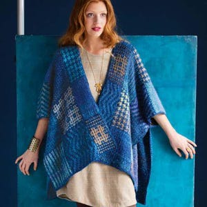 Puede incluir: Un poncho de punto azul con diseño de patchwork y cuello en V. El poncho presenta varios tonos de azul y blanco. La persona lleva un brazalete dorado y un collar con colgante dorado.
