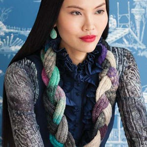 May include: A woman wearing a multi-colored braided knit scarf with a blue, green, purple, and gray color scheme. The scarf is worn around the neck and drapes down the front of the chest.