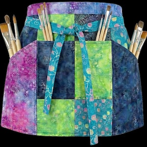 May include: A colorful artist's apron with multiple pockets, filled with paint brushes. The apron features a patchwork design in shades of purple, blue, and green, with a turquoise tie. The apron is designed to hold art supplies.
