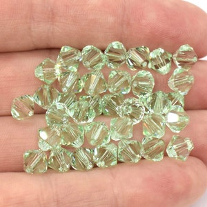 May include: A close-up of a hand holding a pile of light green crystal beads. The beads are faceted and have a shiny finish.