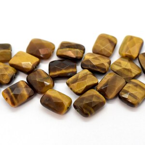May include: A group of 16 faceted tiger's eye beads in a rectangular shape. The beads are a mix of brown and gold colours.