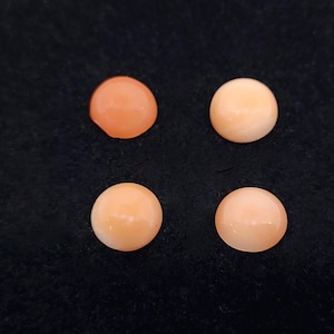 May include: Four round, polished coral cabochons in varying shades of peach and light orange are arranged on a black background. The gemstones have a smooth, glossy surface, and are likely for jewelry making or crafting.