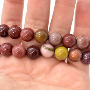 May include: A strand of multi-colored gemstone beads. The beads are round and have a polished finish. The colors of the beads are brown, pink, green, and yellow.