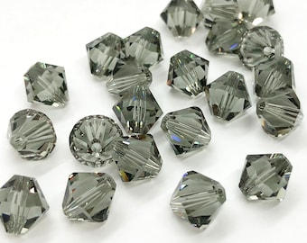 Könnte beinhalten: Eine Gruppe von 20 dunkelgrauen facettierten Glasperlen. Die Perlen sind pyramidenförmig mit quadratischer Basis. Die Perlen sind auf einer weißen Oberfläche verstreut.