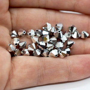 May include: A collection of silver-colored, faceted beads. The beads have a geometric shape with multiple sides, reflecting light. They are small and appear to be made of a metallic material, possibly for jewelry making or crafts.