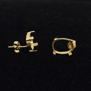 Puede incluir: Dos pendientes de oro. Un pendiente es de estilo poste con un pequeño respaldo redondo y un elemento decorativo. El otro pendiente tiene forma ovalada abierta con un pequeño elemento decorativo. Los pendientes están sobre un fondo negro.