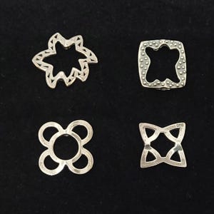 Peut inclure: Quatre breloques en métal argenté aux formes géométriques et organiques distinctes. L'une est en forme d'étoile avec des découpes triangulaires, une est carrée avec des bords pointillés, une est un trèfle à quatre cercles et une est une étoile à quatre branches.