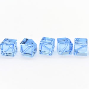 May include: Seven light blue crystal beads in a cube shape with a faceted design. The beads are arranged in a row on a white surface.