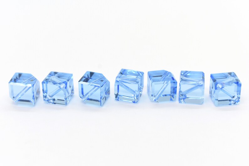 May include: Seven light blue crystal beads in a cube shape with a faceted design. The beads are arranged in a row on a white surface.