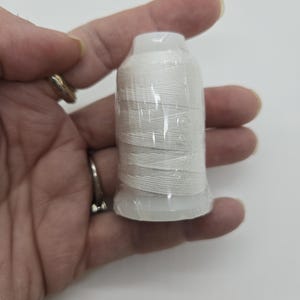 May include: A white thread spool, wrapped in clear plastic, held in a hand. The thread is tightly wound around a white plastic base and top. The background is plain white.
