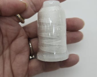 May include: A white spool of thread, wrapped in clear plastic, held in a hand. The thread is tightly wound around a white plastic base and top. The background is a plain white.