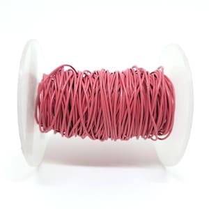 Puede incluir: Un carrete de cordón de cuero rosa. El cordón está enrollado firmemente alrededor de una bobina de plástico blanco. El cordón de cuero es de color rosa polvoriento y parece delgado y flexible. El carrete está sobre un fondo blanco.