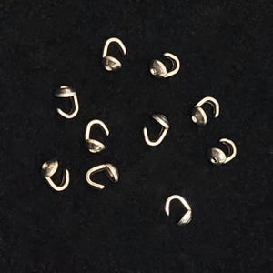 May include: A collection of small, silver-colored crimp bead covers, also known as bead tips, displayed against a dark background. These jewelry findings are designed to conceal crimp beads, providing a clean finish for jewelry making.