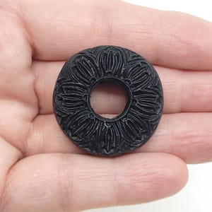 May include: A black, circular object with a floral pattern carved around the outer edge and a hole in the center. The object is held in a hand against a white background. The carved design features detailed petal-like shapes.