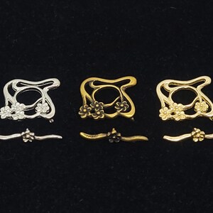 Könnte beinhalten: Drei metallene Blumenringrohlinge in Silber, Gold und Messing. Jeder Rohling hat ein zartes Blumenmuster und eine kreisförmige Öffnung in der Mitte. Die Rohlinge sind in einer Reihe von links nach rechts angeordnet, wobei der silberne Rohling links, der goldene Rohling in der Mitte und der messingfarbene Rohling rechts liegt.