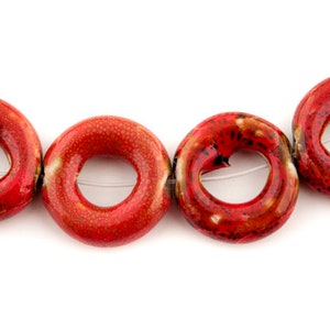 Puede incluir: Una hebra de cuentas de cerámica roja en forma de rosquilla con motas blancas. Las cuentas están enhebradas en un hilo transparente.