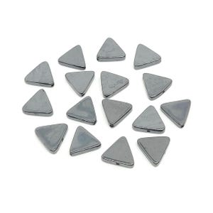 May include: A collection of triangular, grey beads with a metallic sheen. Each bead has a smooth surface and a hole for stringing. The beads are scattered on a white background, suitable for jewellery making or crafting.