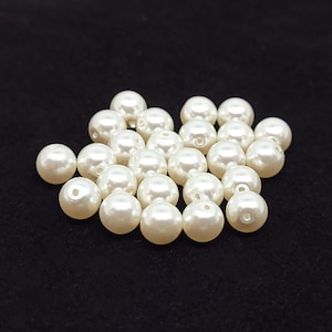 Puede incluir: Una colección de pequeñas cuentas redondas de perlas artificiales en color blanco roto. Cada cuenta presenta una superficie lisa y brillante con un único orificio central para enhebrar. Las cuentas están dispuestas en un grupo, mostrando su forma esférica.