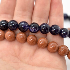 May include: Two strands of beads, one brown and one blue, are shown in a hand. The brown beads are smooth and have a speckled texture. The blue beads are a darker shade of blue and have a speckled texture.