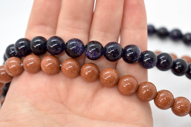 May include: Two strands of beads, one brown and one blue, are shown in a hand. The brown beads are smooth and have a speckled texture. The blue beads are a darker shade of blue and have a speckled texture.