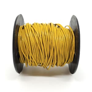 Puede incluir: Un carrete de cordón de cuero amarillo. El cordón está enrollado firmemente alrededor de un carrete de madera gris oscuro. El cordón parece liso y tiene un ligero brillo. El carrete está centrado sobre un fondo blanco.