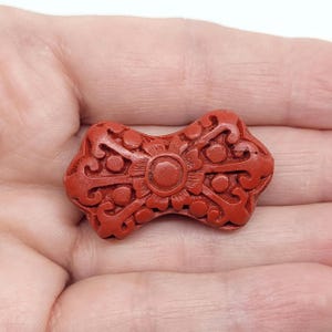 May include: A carved red decorative object with an intricate floral design. The object is held in a hand and has a central circular element surrounded by detailed patterns. The color is a deep, rich red.