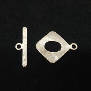 May include: A silver-colored toggle clasp set, featuring a bar and a diamond-shaped component with an oval opening. The clasp has a textured surface and small loops for attachment. Ideal for jewelry making.