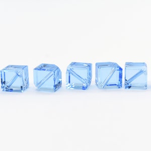 May include: Seven light blue glass cube beads with a clear, faceted design. The beads are arranged in a row on a white background.