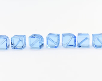 May include: Seven light blue glass cube beads with a clear, faceted design. The beads are arranged in a row on a white background.