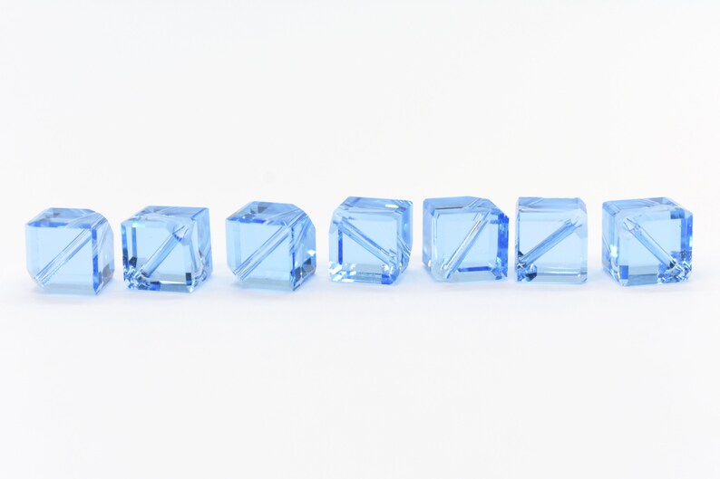 May include: Seven light blue glass cube beads with a clear, faceted design. The beads are arranged in a row on a white background.