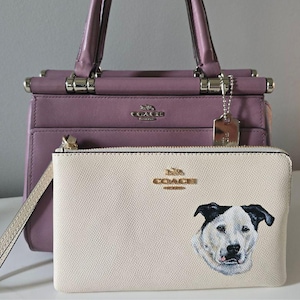 May include: A lilac handbag with silver hardware and a matching wristlet. The wristlet features cream leather with a dog portrait. Both items have the Coach logo.