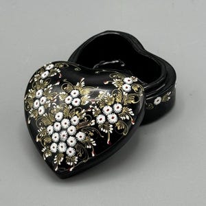 Könnte beinhalten: Eine schwarze herzförmige Schmuckschatulle mit einem floralen Design, das in Weiß und Gold bemalt ist. Der Deckel ist geöffnet und zeigt das schwarze Innere.
