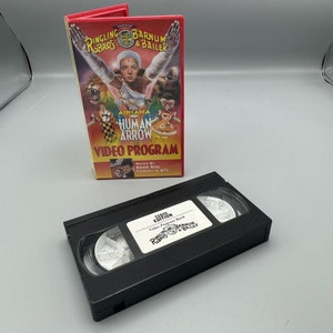 Peut inclure: Une cassette VHS noire avec une étiquette rouge et blanche qui indique "120th Edition Video Program Book Ringling Bros. Barnum & Bailey". L'étiquette présente également un logo de clown avec un nez rouge et un chapeau rayé rouge et blanc.
