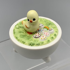 May include: A white ceramic figurine of a yellow chick sitting on a bed of green grass with colorful Easter eggs. The figurine is on a white base with three legs.