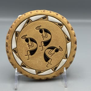 May include: Round, tan-colored ceramic coaster featuring three black Kokopelli figures in the center. The coaster has a white and black geometric border and a textured edge. The coaster is displayed on a clear stand.