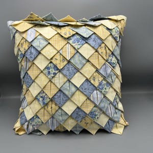 May include: A square decorative pillow with a patchwork design. The pillow features a repeating pattern of yellow, blue, and white fabric squares. Some squares have floral patterns, while others are solid or checkered.