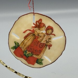 May include: A round, cream-colored Christmas ornament with a red border. The ornament features a vintage illustration of two carolers in red and gold attire, holding a songbook. A large red bow and green holly complete the festive design.