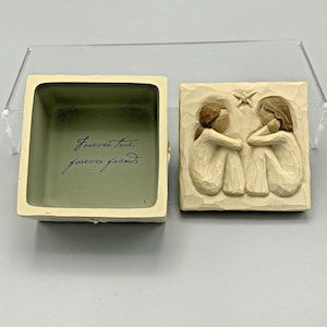 Könnte beinhalten: Eine kleine, quadratische, beige Box mit einem geschnitzten Holzdeckel. Der Deckel zeigt zwei Engelfiguren, die sich in den Armen liegen. Das Innere der Box ist beige und trägt die Inschrift "Forever true, forever friends".