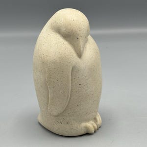 May include: A speckled, off-white penguin ornament. The penguin is in a curved pose, with its head tucked down and its wings close to its body. The sculpture has a smooth, rounded form and a matte finish.