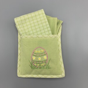 May include: Green fabric Easter egg holder with a green and white plaid fabric lining. The holder has an embroidered Easter egg with yellow and pink details.