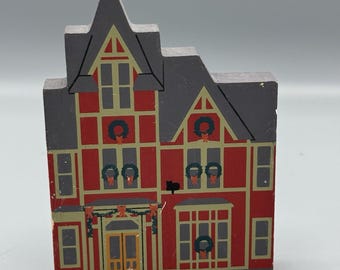 May include: A wooden decorative house with a red facade, gray roof, and gold trim. The house features windows, wreaths, and a door. The design is reminiscent of a festive holiday theme.