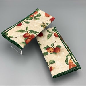 Peut inclure: Deux serviettes en tissu de couleur crème avec une bordure verte et un motif de pommes, de poires et de feuilles rouges et orange.