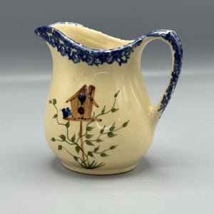May include: Cream-colored ceramic pitcher with a blue floral rim and handle. The pitcher features a hand-painted birdhouse design with green vines and blue flowers. A decorative piece for the kitchen or dining room.