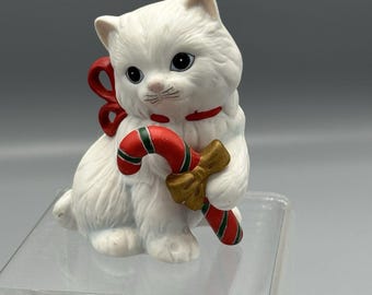 May include: A white ceramic cat figurine with a red ribbon and a candy cane. The cat is holding a red and green candy cane with a gold bow. The figurine has black eyes and a pink nose.
