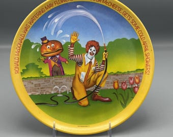 May include: A yellow plastic plate with a cartoon image of Ronald McDonald watering flowers while Mayor McCheese gets wet. The text on the plate reads "Ronald McDonald was watering may flowers... but Mayor McCheese gets some cold April showers!"